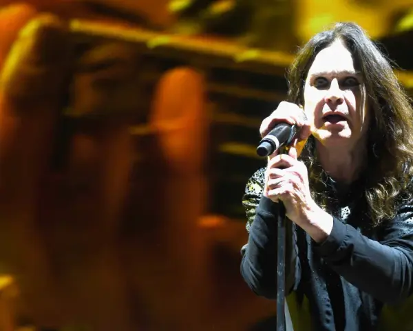 Ozzy Osbourne śpiewa do mikrofonu na koncercie, z rozmytym tłem w kolorach żółci i pomarańczy.