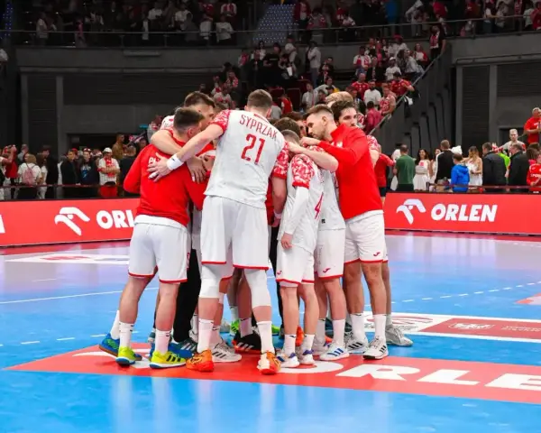Drużyna piłkarzy ręcznych w białych strojach z czerwonymi elementami tworzy zgraną grupę na boisku.