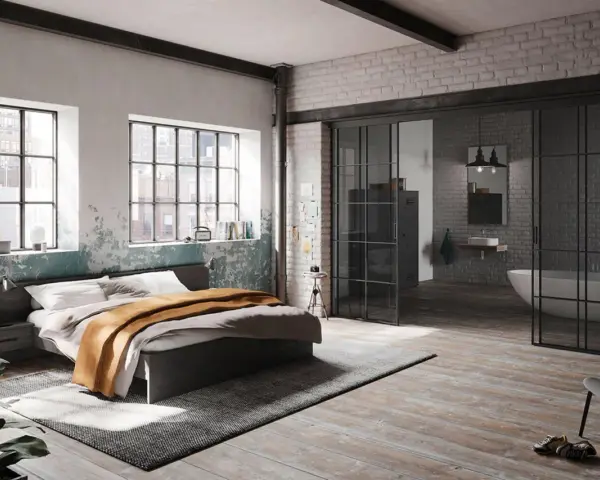 Ein gemütliches **Bett unter Fenster** mit industriellem Charme. Daneben ein offener Badbereich mit Wanne.