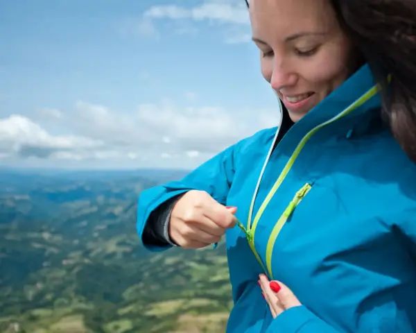 Donna con impermeabile montagna blu, chiude la zip. Paesaggio verdeggiante sotto un cielo azzurro.