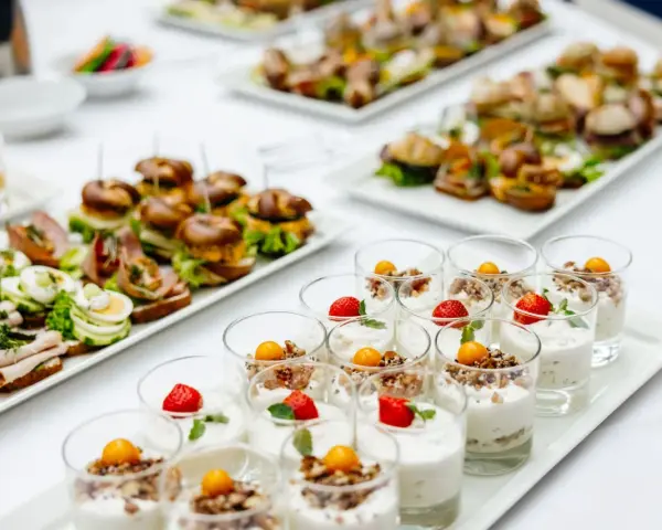 Varietà di antipasti freddi per buffet: mini hamburger, tartine con uova e salumi, e dolcetti in bicchiere con frutta e yogurt.