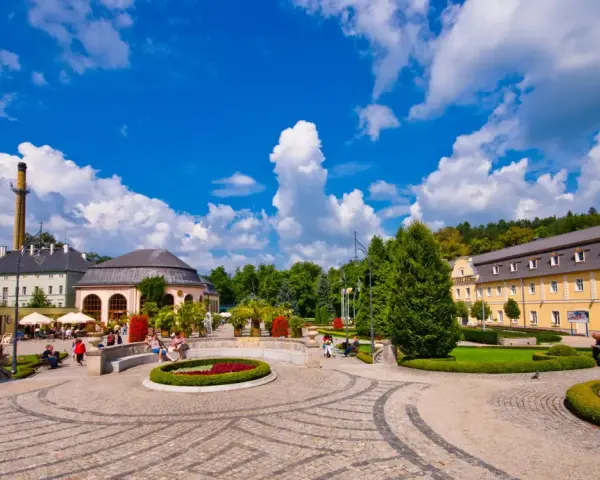 Słoneczny dzień w Kudowie Zdrój. Spacerowicze odpoczywają w pięknym parku, podziwiając architekturę i zieleń. To idealne miejsce na relaks i odkrywanie atrakcji.