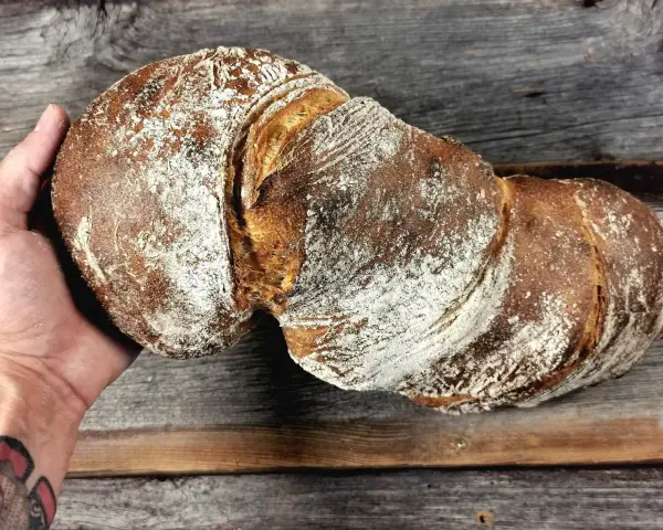Ein rustikales wurzelbrot mit sauerteig, bestäubt mit Mehl, liegt auf einem Holzbrett.