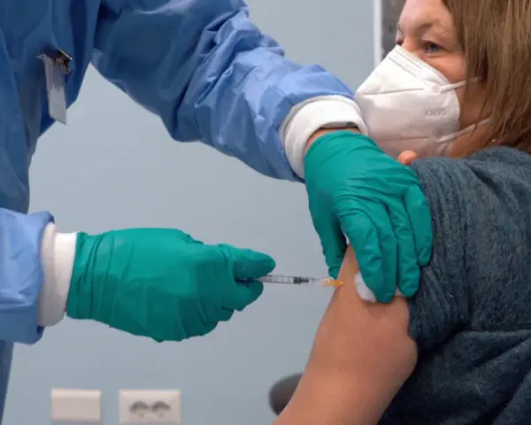 Osoba w niebieskim fartuchu i zielonych rękawiczkach podaje zastrzyk. Kobieta w masce KN95 otrzymuje trzecią dawkę szczepionki.