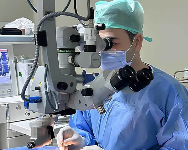 Chirurgo con maschera e camice blu opera con microscopio Zeiss. Monitor vitale e strumentazione indicano un intervento post-operatorio vitrectomia gas.