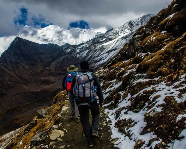 Dwóch turystów z plecakami wędruje ścieżką u podnóża ośnieżonych gór, przypominających krajobraz Mount Kenya.