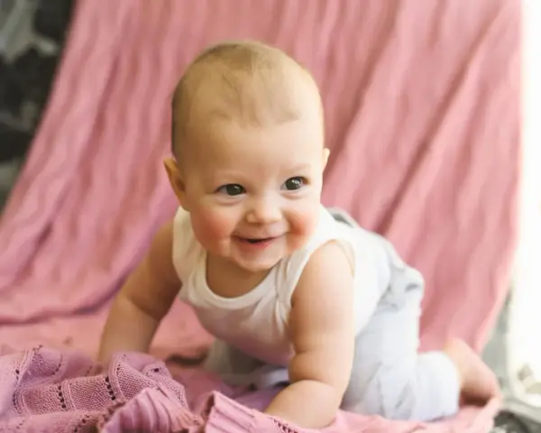 Ein lächelndes Baby krabbelt auf einer rosafarbenen Decke. Es ist etwa 26 Wochen alt und erlebt gerade einen Entwicklungsschub.