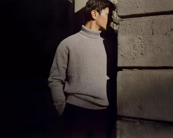 Uomo con un elegante outfit maglione collo alto grigio, in piedi vicino a un muro di pietra.