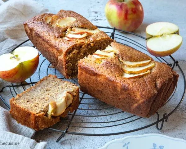 Saftiger Apfelkuchen mit Apfelmus-Note, dekoriert mit frischen Apfelscheiben. Daneben liegen Äpfel und ein Stück Kuchen.