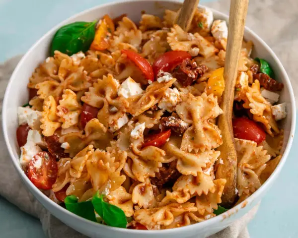 Ein köstlicher, sommerlicher Nudelsalat mit Farfalle, Tomaten, Feta und Basilikum, serviert in einer weißen Schüssel mit Holzutensilien.
