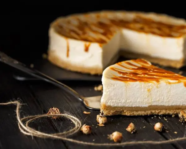 Una fetta di cheesecake senza panna, con glassa caramellata, su un piatto da portata, con la torta intera sullo sfondo.
