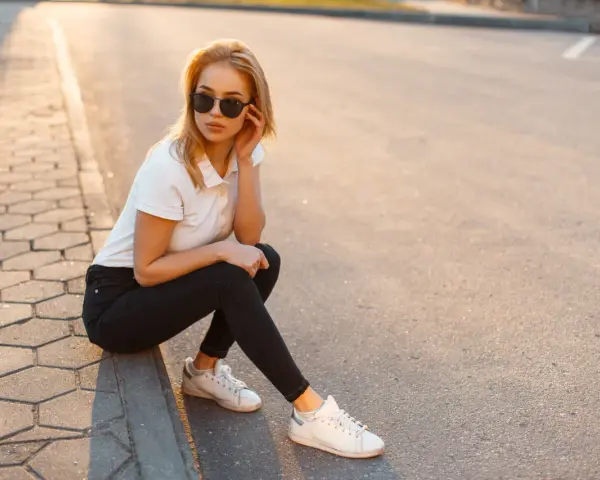 Blondynka w czarnych jeansach i białej koszulce polo, siedząca na chodniku. Stylowa inspiracja, jak nosić koszulka polo damska na co dzień.