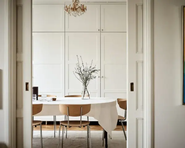 Comedor con mesa cubierta de mantel blanco, sillas de madera y un gran armario empotrado. El estilo ncs blanco aporta luminosidad.