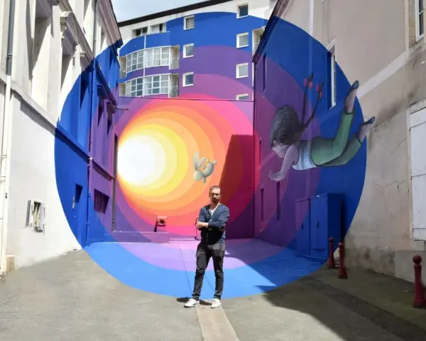 Un artiste street art français pose devant sa fresque colorée d'une fillette et d'un lapin flottant dans un tunnel psychédélique.