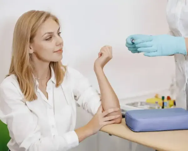 Kobieta w białej koszuli przygotowuje się do pobrania krwi. Pielęgniarka w rękawiczkach trzyma probówkę. To standardowa procedura, gdy badanie wykonuje się na czczo.
