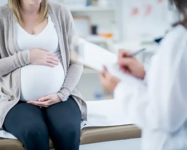 Kobieta w ciąży siedzi na kozetce, trzymając dłonie na brzuchu. Lekarz spisuje dokumenty, prawdopodobnie wystawiając zaświadczenie o ciąży.