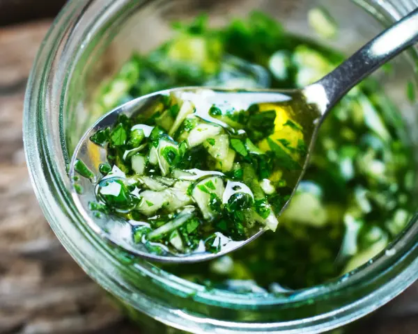 Łyżka z aromatycznym, zielonym sosem chimichurri, pełnym ziół i czosnku, zanurzona w słoiku.
