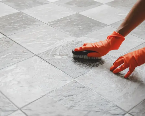 Mani con guanti arancioni puliscono le fughe del pavimento con una spazzola, mostrando come pulire le fughe del pavimento.