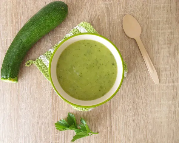 Zucchini Brei Baby: Eine Schüssel grüner Zucchinipüree, daneben eine Zucchini und ein Holzlöffel.