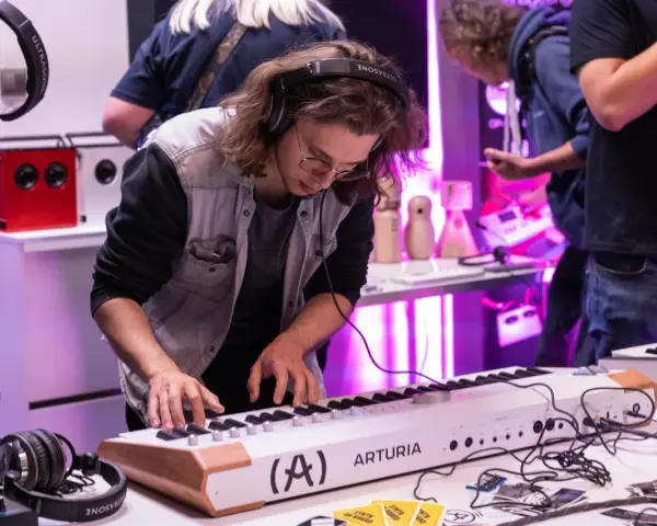 Młody mężczyzna w okularach i słuchawkach ćwiczy na keyboardzie Arturia. To część kursu produkcji muzycznej, gdzie można poznać tajniki tworzenia dźwięków.