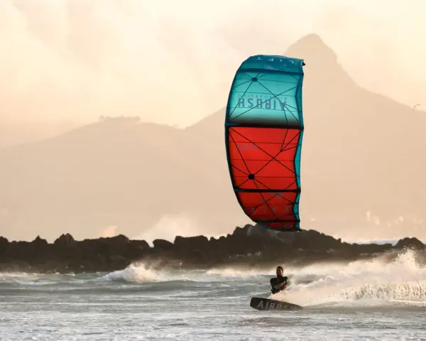 Kitesurfer na desce w akcji, z kolorowym latawcem na tle gór i morza.