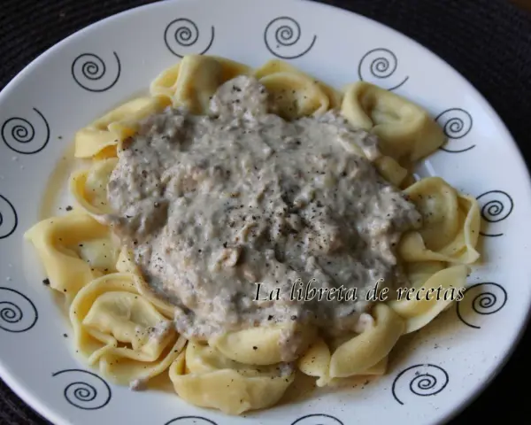 Tortellini de queso cubiertos con una cremosa salsa de champiñones y pimienta negra, servidos en un plato blanco con espirales decorativas.