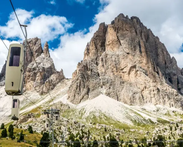 Cabine della funivia salgono verso le imponenti vette del Sassolungo, un anello di roccia maestoso sotto un cielo azzurro con nuvole bianche.