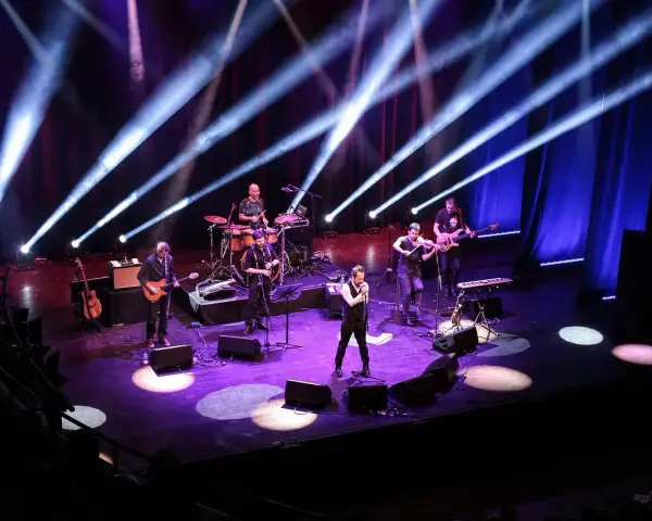 Zespół muzyczny na scenie, oświetlony niebieskimi i fioletowymi światłami. Wokalista na pierwszym planie, gitarzyści, basista, skrzypek i perkusista. Jak liczyć takty w muzyce, można się nauczyć, obserwując ich rytm.
