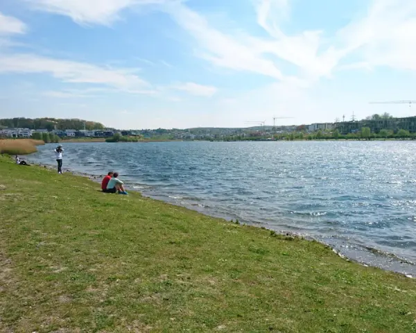Ein sonniger Tag am Dortmunder See. Menschen spazieren am Ufer entlang, während ein Paar auf dem Gras sitzt und die Aussicht genießt.