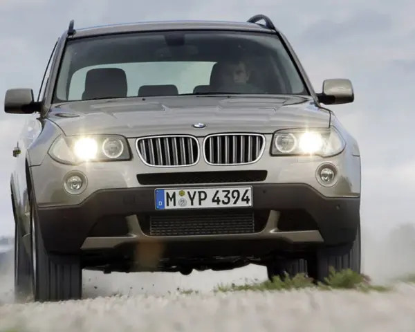 Złoty SUV BMW X3 z włączonymi światłami jedzie po szutrowej drodze, unosząc kurz. To jeden z najbardziej komfortowych aut na drodze.