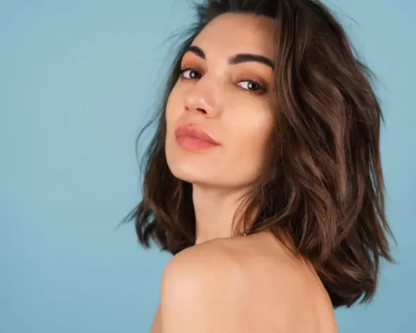 Kobieta z lekko cieniowanymi włosami do ramion, zmysłowo spogląda przez ramię na błękitnym tle.