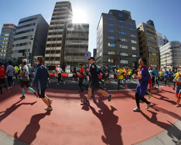 Atleti corrono nella maratona di Tokyo, circondati da grattacieli moderni sotto un sole splendente.