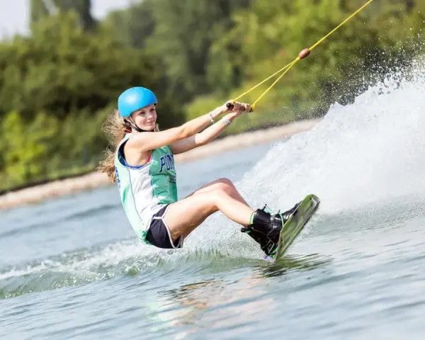 Dziewczyna w kasku i kamizelce pływa na wakeboardzie, tworząc wielki szelment wody.