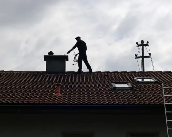 Kominiarz na dachu sprawdza komin. Zastanawiasz się, ile kosztuje przegląd kominiarski?