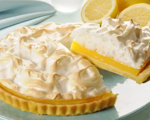 Pyszna tarta cytrynowa z bezą, z kawałkiem na łopatce. Obok leżą połówki cytryn.