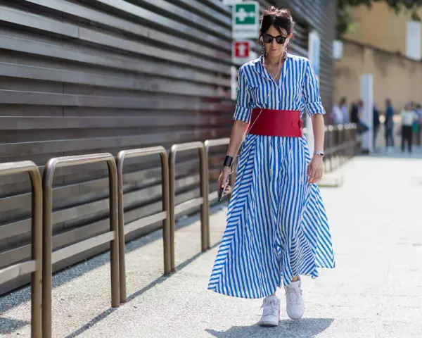 Donna con vestito a righe blu e bianche, cintura rossa e sneakers bianche cammina per strada.