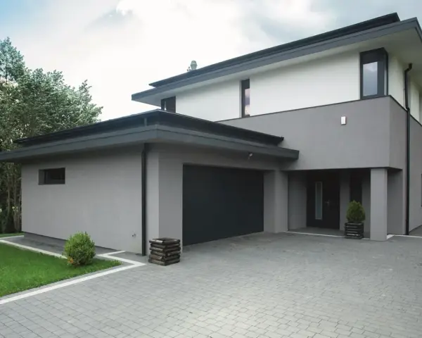 Nowoczesny dom z szarymi i białymi tynkami, z ciemnym dachem i bramą garażową. Eleganckie kolory tynków na domach tworzą minimalistyczną całość.