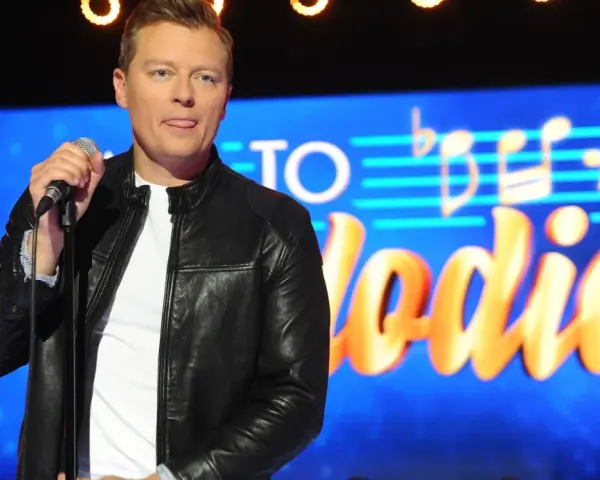 Rafał Brzozowski z mikrofonem na scenie, w tle napis "Jaka to melodia?".