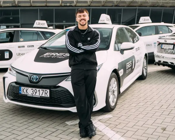 Młody mężczyzna w dresie stoi przed białymi samochodami Uber z napisem "ERES Partner". To jego praca prywatny kierowca.