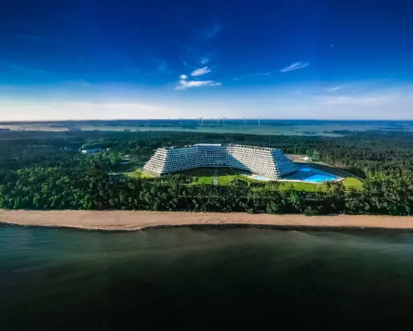 Hotel Gołębiewski w Pobierowie, otoczony lasem, z basenem i widokiem na morze. W oddali widać farmę wiatrową.