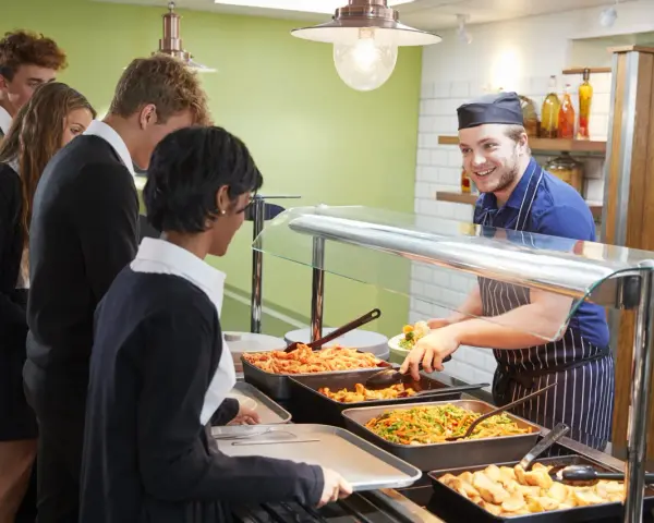 Des élèves font la queue pour le déjeuner à la cantine. Un employé souriant sert des légumes. Un contrat extra restauration est en cours de discussion.