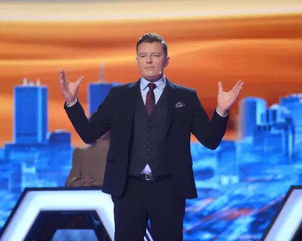 Rafał Brzozowski w garniturze, z rękami uniesionymi na boki, prowadzi program "Jaka to melodia?".