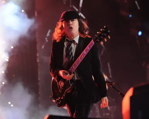 Ein Mann mit Hut und Schlagzeug spielt auf einer ACDC Gitarre, umgeben von Rauch und Lichtern.