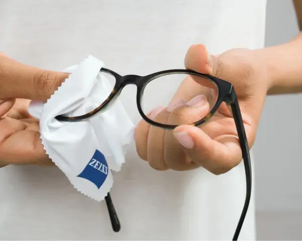 Här putsas glasögon med en mjuk trasa. En person håller i bågarna och putsar linserna med en vit trasa med Zeiss-logga.