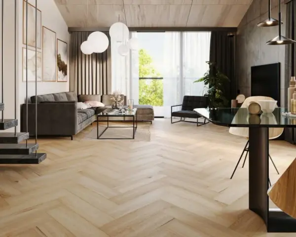 Nowoczesny salon z szarą sofą, szklanym stolikiem i jadalnią. Wzrok przyciągają panele bez dylatacji w progu, tworzące jednolitą, drewnianą podłogę w jodełkę.