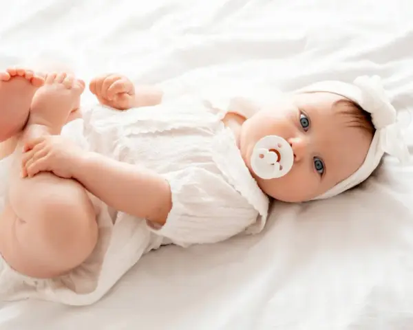 Bebé Ariadna, con su chupete y diadema blanca, juega con sus pies sobre una cama blanca.
