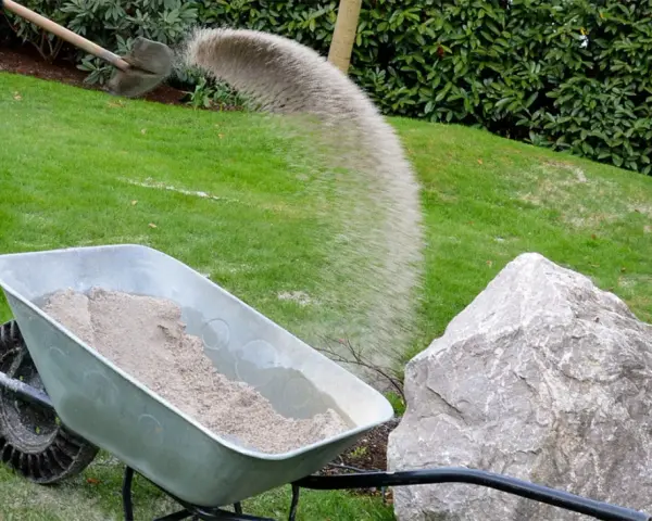Schubkarre mit Sand, während eine Schaufel Sand auf den Rasen rieselt.