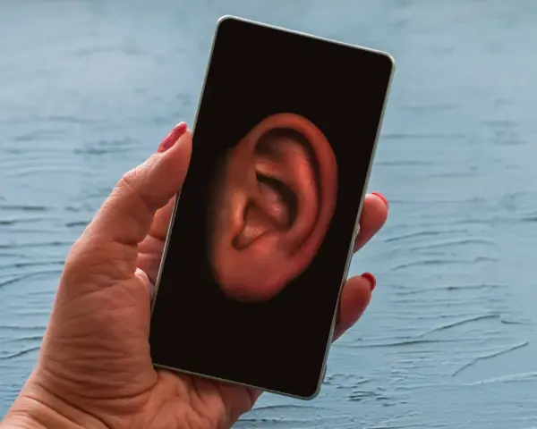 Dłoń trzyma smartfon z uchem na ekranie. Czy Twój telefon jest na podsłuchu? Sprawdź to!