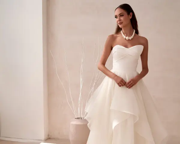Elegancka panna młoda w sukni ślubnej z falbanami, prezentująca najmodniejsze suknie ślubne. Delikatny naszyjnik z pereł podkreśla jej urodę.