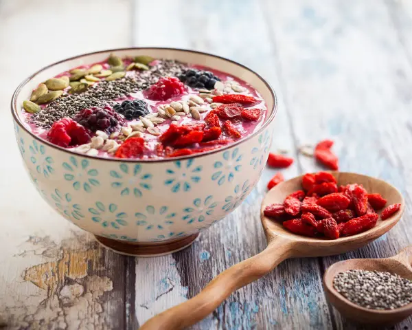 Miseczka smoothie z owocami, nasionami chia, pestkami dyni i słonecznika. Obok łyżki z jagodami goji i nasionami chia. Pyszny sposób, jak jeść nasiona chia.